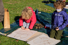 Two children sitting on the ground drawing with walnuts on brown paper.